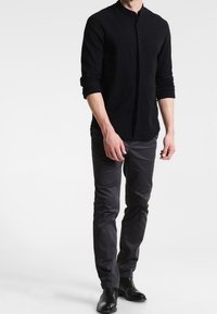 Uomo che indossa una camicia nera a colletto mandarina, pantaloni slim-fit grigio scuro e stivali neri in pelle, in piedi contro uno sfondo bianco.