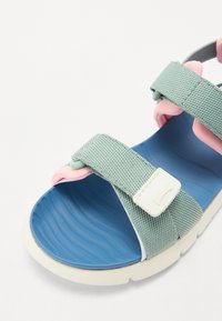 Sandal de tecido verde com detalhes em rosa, tiras ajustáveis, parte superior texturizada e palmilha azul almofadada; sola de borracha branca com padrão de ondas.