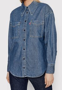 Chemise en denim bleu foncé avec un col pointu, des boutons sur le devant, deux poches poitrine et des détails de couture. Les manches longues sont dotées de poignets à boutons.