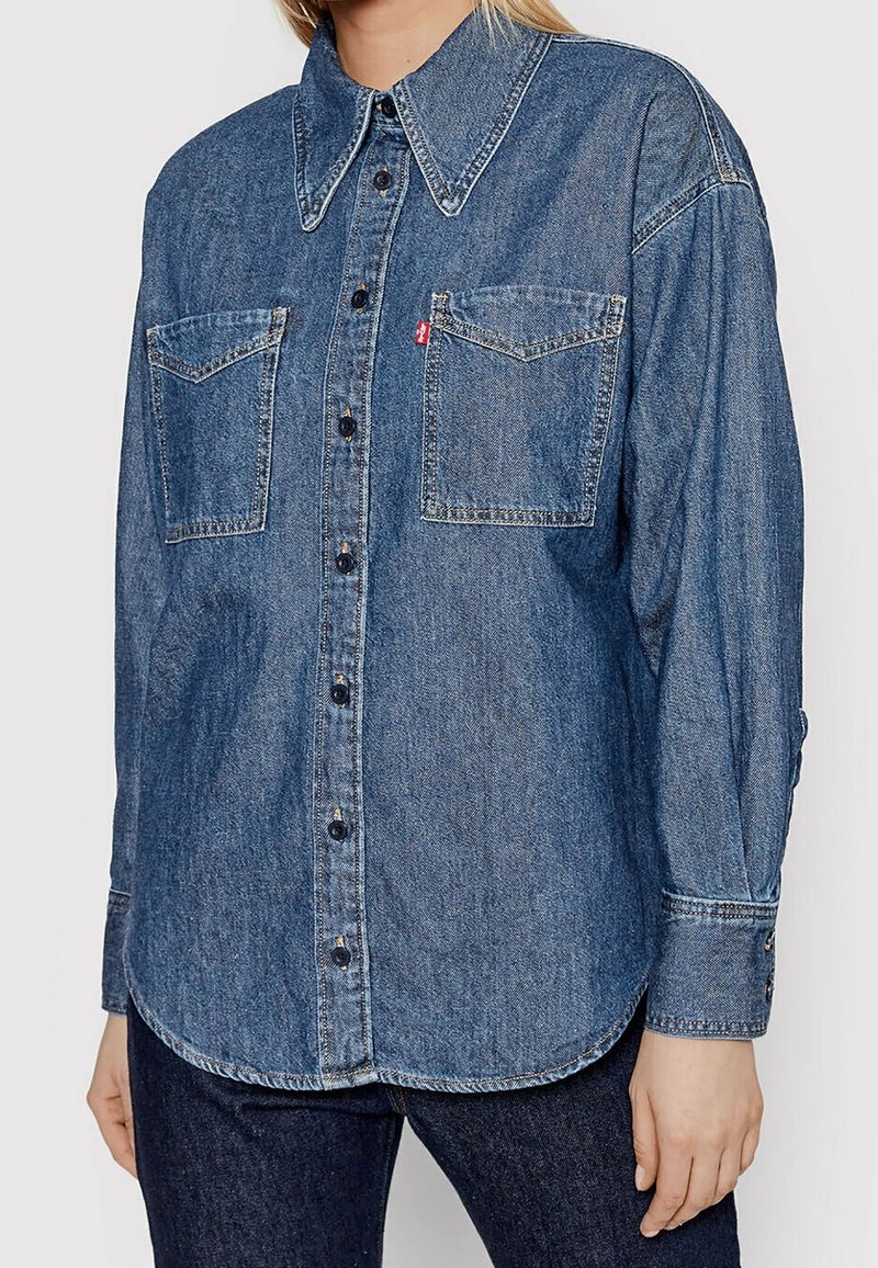 Chemise en denim bleu foncé avec un col pointu, des boutons sur le devant, deux poches poitrine et des détails de couture. Les manches longues sont dotées de poignets à boutons.
