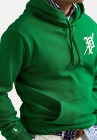 Grön hoodie av mjuk bomull med en framficka. Har en vit logotyp på bröstet och ribbade muddar. Lager-på-lager över en randig tröja.