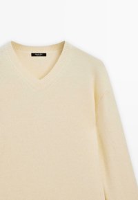 Pull-over beige en V, fabriqué en maille douce. Présente un col et des coutures côtelés, avec des manches longues et une coupe décontractée.