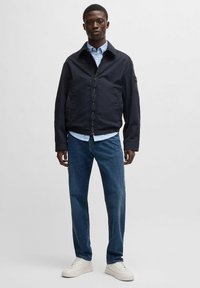 Marinblå bomberjacka med dragkedja fram, sidofickor och uppvikt krage, i kombination med en ljusblå skjorta, blå jeans och vita sneakers.