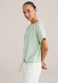 Hele rohelise, lühikeste varrukatega pluus, mis on valmistatud pehmest kangast ja millel on sõlm üksikasjades taljes. Koos valgete teksadega, sujuva tekstuuriga.