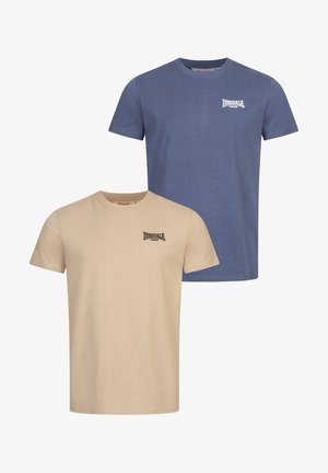 Beige pamut póló, bal mellen egy kis fekete logóval, fekete nadrággal és fehér sportcipővel párosítva, semleges háttér előtt.