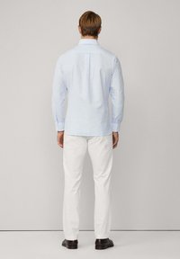 Lichtblauwe overhemd met lange mouwen en een knoopsluiting kraag, rugplooien, en witte katoenen broek, gedragen met donkere schoenen, tegen een neutrale achtergrond.