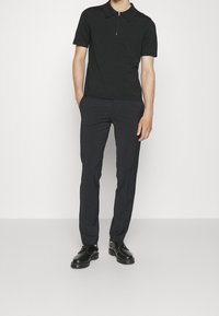 Černé polo tričko s krátkým rukávem a zipovým límcem, spárované s tmavými slim fit kalhotami a černými šněrovacími botami na jednolitém pozadí.