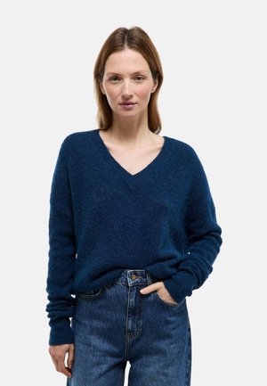 Femme aux cheveux châtain clair portant un pull bleu foncé à col en V et un jean bleu, debout avec une main dans la poche, sur fond blanc.