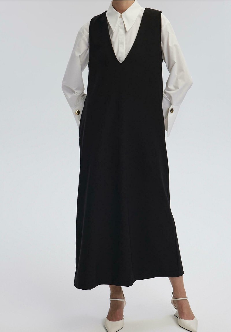 Personne portant une longue robe noire sans manches par-dessus une chemise blanche avec des boutons de manchette dorés et des chaussures blanches à talons pointus.