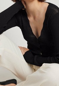 Schwarze, gerippte Langarm-Cardigan mit tiefem V-Ausschnitt und Knopfverschluss, ausgestattet mit einem figurbetonten Design und strukturiertem Stoff. Getragen mit weißen Hosen.