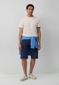Lichtbeige T-shirt met ronde hals, marineblauwe cargo-shorts, blauwe sweatshirt geknoopt om de taille, en witte sneakers met beige accenten.