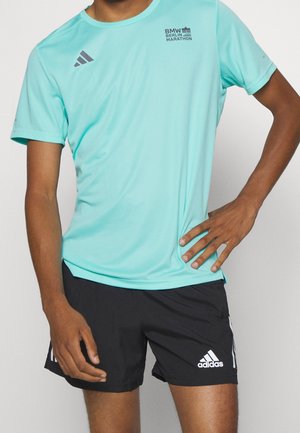 Mężczyzna noszący jasno niebieską koszulkę BMW Berlin Marathon i czarne spodenki Adidas z białym logo i paskami, pozujący z ręką na biodrze.