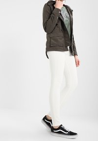 Veste zippée olive avec deux poches avant, associée à un t-shirt graphique vert clair, un jean skinny blanc et des baskets noires.