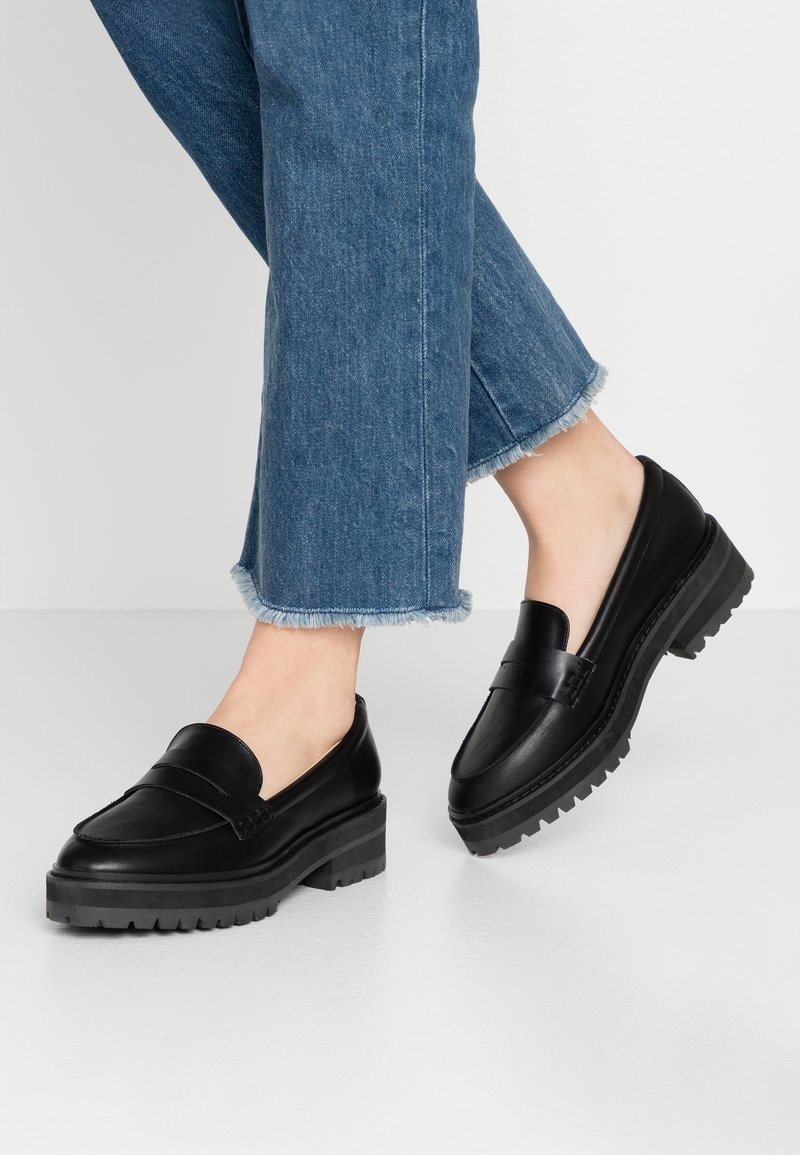 Jambes portant un jean court bleu avec des bords effilochés et des mocassins en cuir noirs à semelles épaisses sur un sol clair.