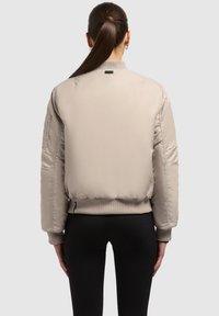 Beige Bomberjacke mit glatter Textur, Rippstrickbündchen und -saum sowie einer Reißverschlusstasche am linken Ärmel. Rückansicht abgebildet.