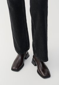 Brown leather ankle boots with a square toe and block heel, paired with black wide-leg jeans. Smooth texture and minimalistic design.