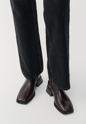 Bottines en cuir marron avec un orteil carré et un talon carré, associées à un jeans noir à jambes larges. Texture lisse et design minimaliste.