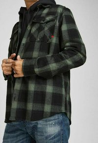 Camicia di flanella a scacchi nera e verde con due tasche frontali, stile con bottoni, indossata sopra un hoodie nero, abbinata a jeans di denim.
