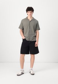Camisa de botón de manga corta, de color verde claro y con una superficie texturizada, combinada con pantalones cortos de mezclilla negros y zapatos grises con rayas negras.