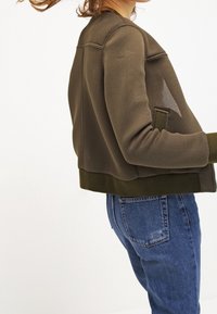 Zielona siatkowa kurtka w oliwkowym kolorze o dopasowanym kroju, z prążkowanym dołem i boczną kieszenią; zestawiona z niebieskimi dżinsami.