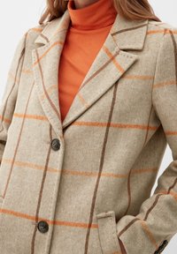 Abrigo de cuadros beige con patrón a cuadros naranja, hecho de tela texturizada. Tiene un cuello afilado y dos botones oscuros en la parte delantera.