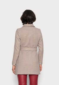 Beige ullkappa med bälte i midjan, slagkrage och långa ärmar. Texturen verkar slät och kompletterar de figursydda röda byxorna.