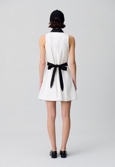 Vestido blanco sin mangas con un cuello negro y un lazo a juego en la cintura, combinado con zapatos negros. Tejido suave y diseño acampanado.