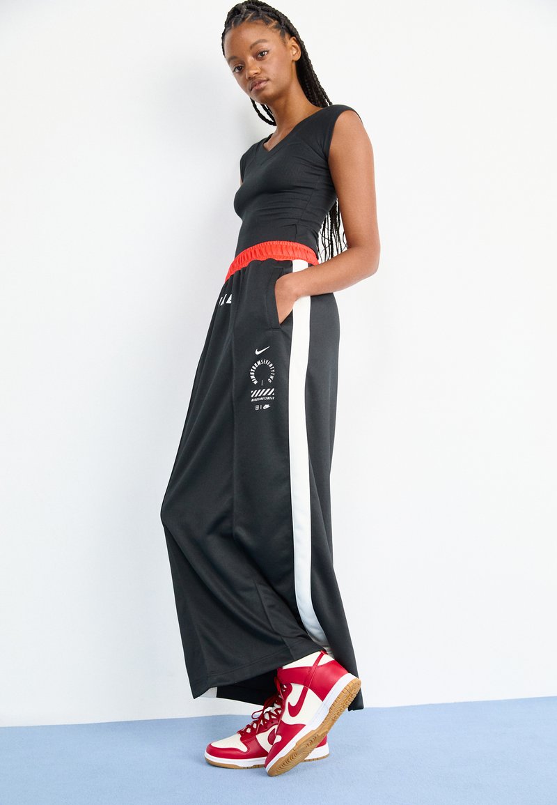 Haut de sport noir et pantalon large noir avec une ceinture élastique rouge. Présente des bandes latérales blanches et un graphisme de marque. Baskets montantes rouges et blanches.