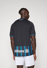 Camisa atlética preta com um painel inferior riscado a azul, apresentando o texto "Allianz" e "No Sweat No Glory" na parte de trás do pescoço. Mangas curtas.