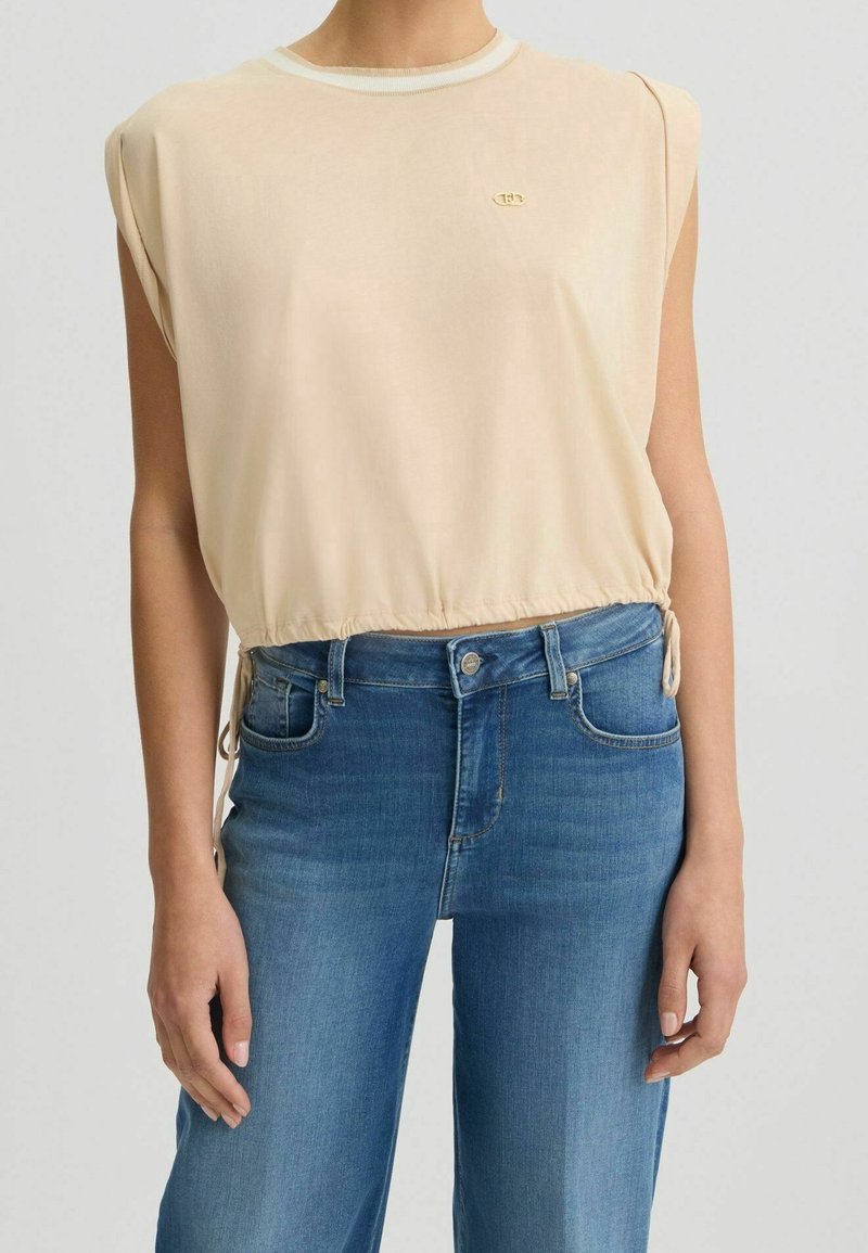 Haut beige sans manches avec légère épaulette et taille à cordon, associé à un jean en denim bleu taille haute.