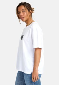 T-shirt bianco in cotone, con maniche corte e una piccola grafica nera sul petto. Abbinato a jeans in denim blu.
