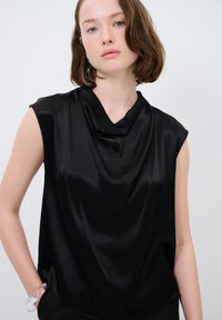 Top senza maniche in satin nero con design a collo a cascata, texture liscia e vestibilità ampia, caratterizzato da un sottile drappeggio nella zona del collo.