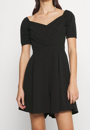 Frau trägt ein schwarzes, kurzärmeliges Kleid mit weitem V-Ausschnitt und plissiertem Rock, accessoriert mit langen hängenden Ohrringen.