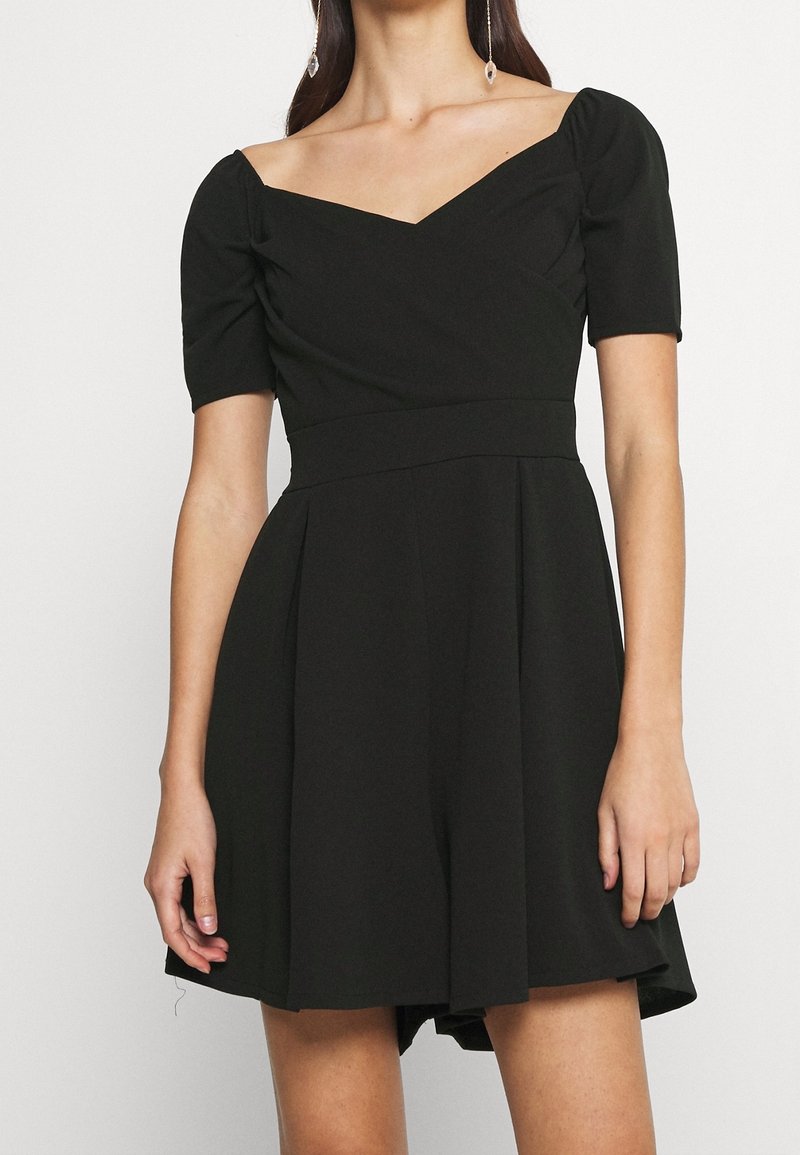 Femme portant une robe noire à manches courtes avec un large décolleté en V et une jupe plissée, accessoirisée avec de longues boucles d'oreilles pendantes.