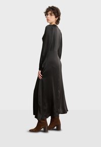 Largo vestido de satén negro con mangas largas, cuerpo ajustado, falda fluida y longitud hasta la mitad de la pantorrilla, combinado con botas de tobillo de ante marrón.