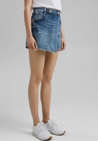 Denim-Minirock mit ausgewaschenem Blauton, fransigem Saum und Vorderseiten-Taschen, kombiniert mit einem lässigen weißen T-Shirt und weißen Turnschuhen.