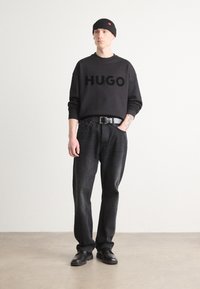 Black sweatshirt with "HUGO" printed prominently, black jeans, and black leather shoes. Model wears a black beanie. Simple, casual outfit.