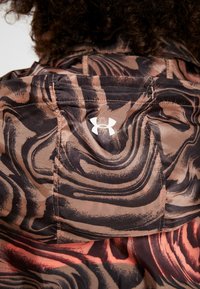 Färgglad jacka i brunt och rosa med mönster, med en slät textur och en vit logotyp. Designen innehåller svängande former och synliga sömmar.