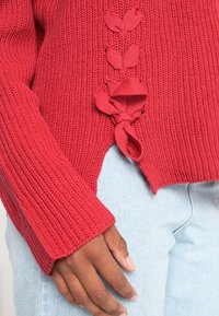 Punainen neulottu neulepaita, jossa on teksturoitu kangas, kukkakoristeiset yksityiskohdat ja sivusaumoissa nauhat. Pitkät hihat, joissa resorit hihansuissa.