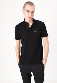Juodas polo marškinėlis, pagamintas iš medvilnės, su trumpu apykakle, trimis sagomis, prigludusia forma ir maža logotipo etikete.