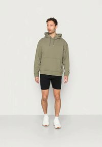 Vīrietis, tērpies olīvzaļā kapucē, melnās šortās, baltās zeķēs un baltās kedās, stāv pretim gaišai fona sērijai un skatās pa kreisi.