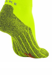 Neon-gelbe Socke mit einem strukturierten Grip-Design, das dezente graue Muster und ein rotes Logo an der Seite zeigt. Leicht, mittelhohe Höhe bis zur Wade.