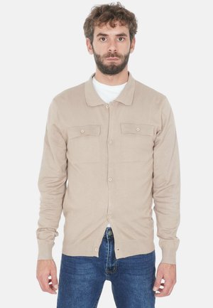 Uomo con barba che indossa un cardigan beige con bottoni sopra una camicia bianca e jeans blu, in piedi contro uno sfondo bianco.