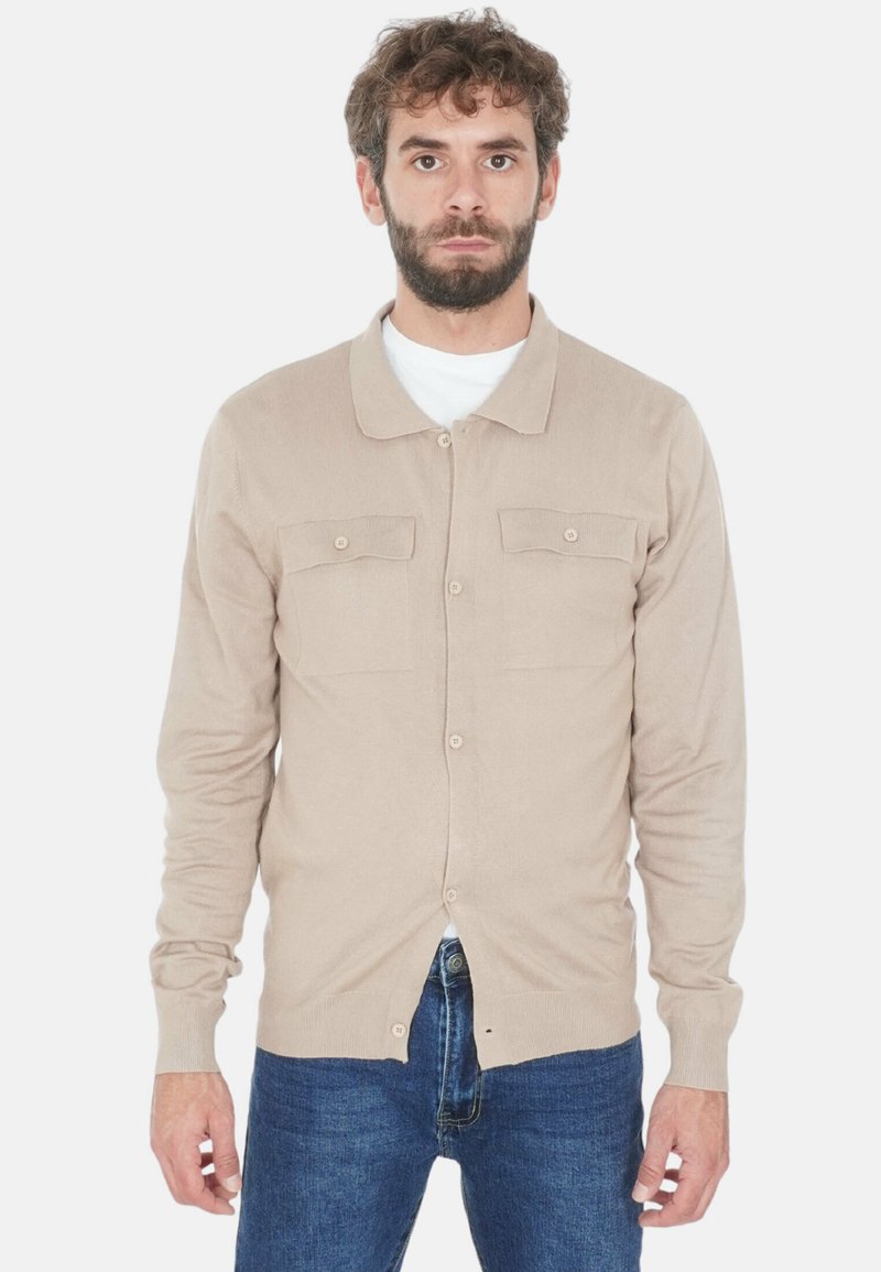 Uomo con barba che indossa un cardigan beige con bottoni sopra una camicia bianca e jeans blu, in piedi contro uno sfondo bianco.