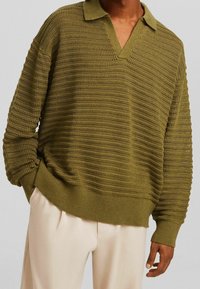 Personne portant un pull en maille texturée vert olive avec col et un pantalon ample de couleur crème, une main dans la poche, sur un fond neutre.