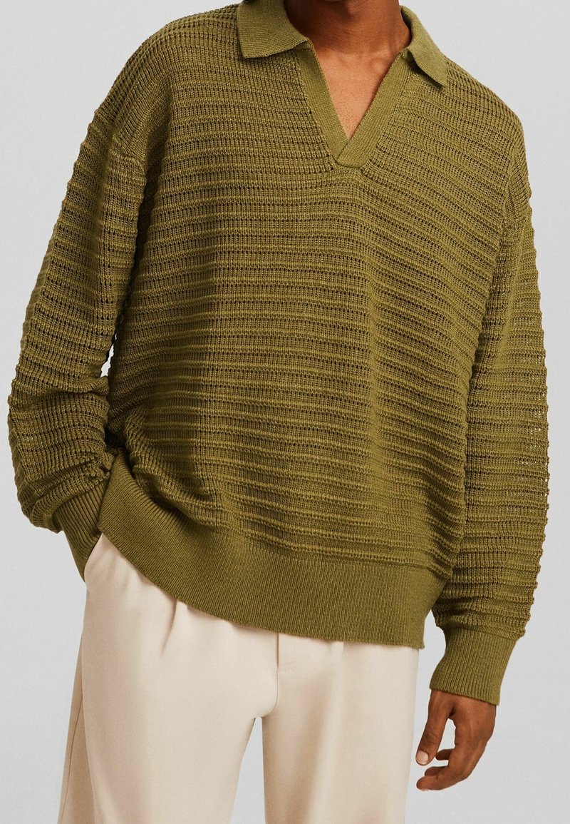 Personne portant un pull en maille texturée vert olive avec col et un pantalon ample de couleur crème, une main dans la poche, sur un fond neutre.