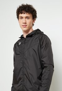 Veste légère zippée noire avec capuche, dotée de poignets élastiques, d'une texture cousue et d'un petit logo sur la poitrine gauche.