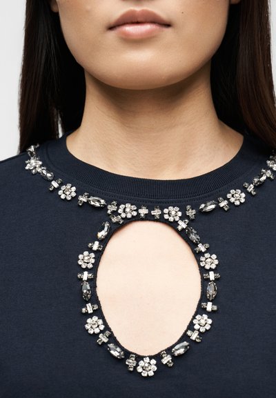 Λαιμός και άνω μέρος στήθους γυναίκας που φοράει μπλε μπλούζα με κοπή σε σχήμα σταγόνας και κοσμήματα γύρω από το λαιμό και την κοπή.