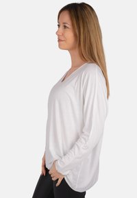Weißes Langarmoberteil mit V-Ausschnitt, weichem Material und lässiger Passform. Die Seiten sind leicht gebogen und der Saum wirkt hinten länger.