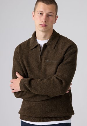 Jeune homme aux cheveux courts portant un pull zippé marron foncé par-dessus une chemise blanche, debout les bras croisés devant un fond gris.